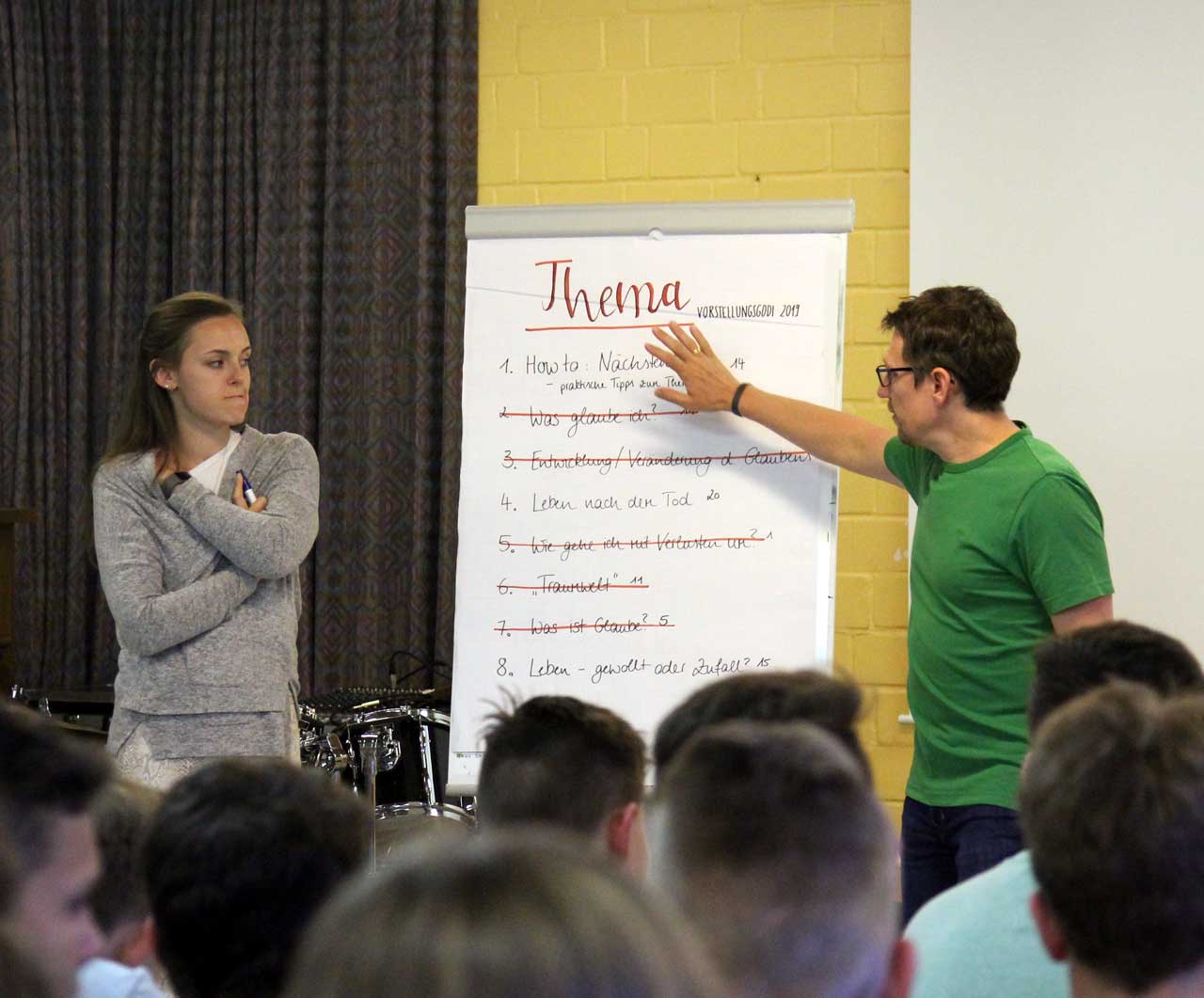 Annika und Thorsten stehen auf neben einem Flipchart. Darauf steht: Thema Vorstellungsgodi 2019. Die Punkte 2,3,5,6 und 7 sind abgestrichen. Auf der Liste stehen noch 1. How to Nächstenliebe, 4. Leben nach dem Tod und 8. Leben - gewollt oder Zufall?.