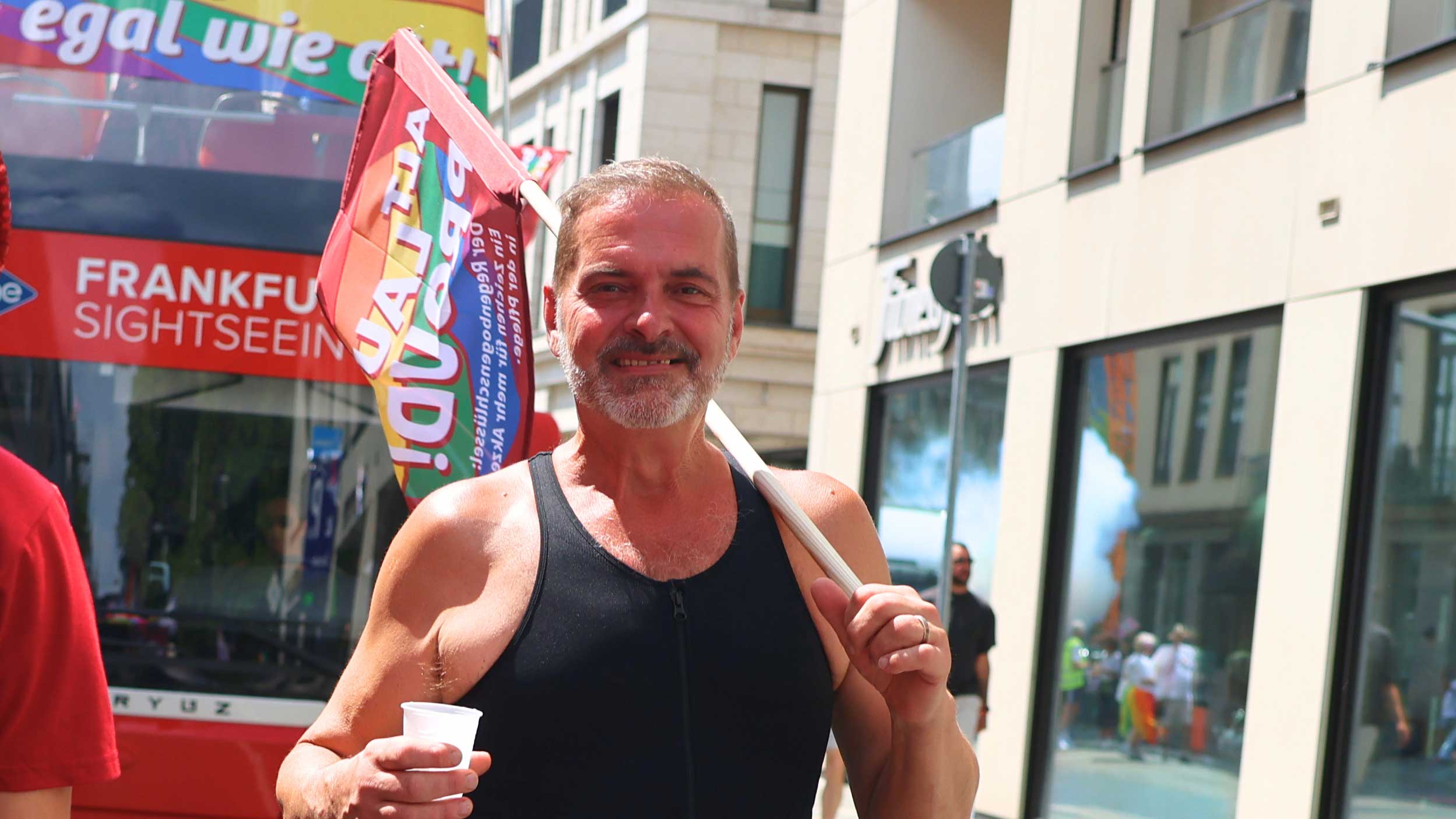 Ein Mann in einem schwarzen Tanktop lächelt, während er einen Becher und eine Regenbogenfahne hält. Er steht im Freien in der Nähe eines roten Sightseeing-Busses und moderner Gebäude.