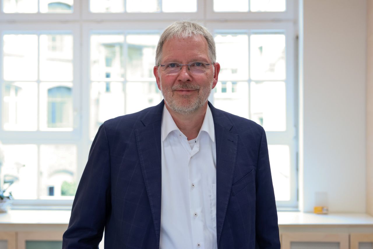 Journalist Arnd Henze steht mit weißem Hemd und blauem Jackett vor einem Fenster und schaut in die Kamera