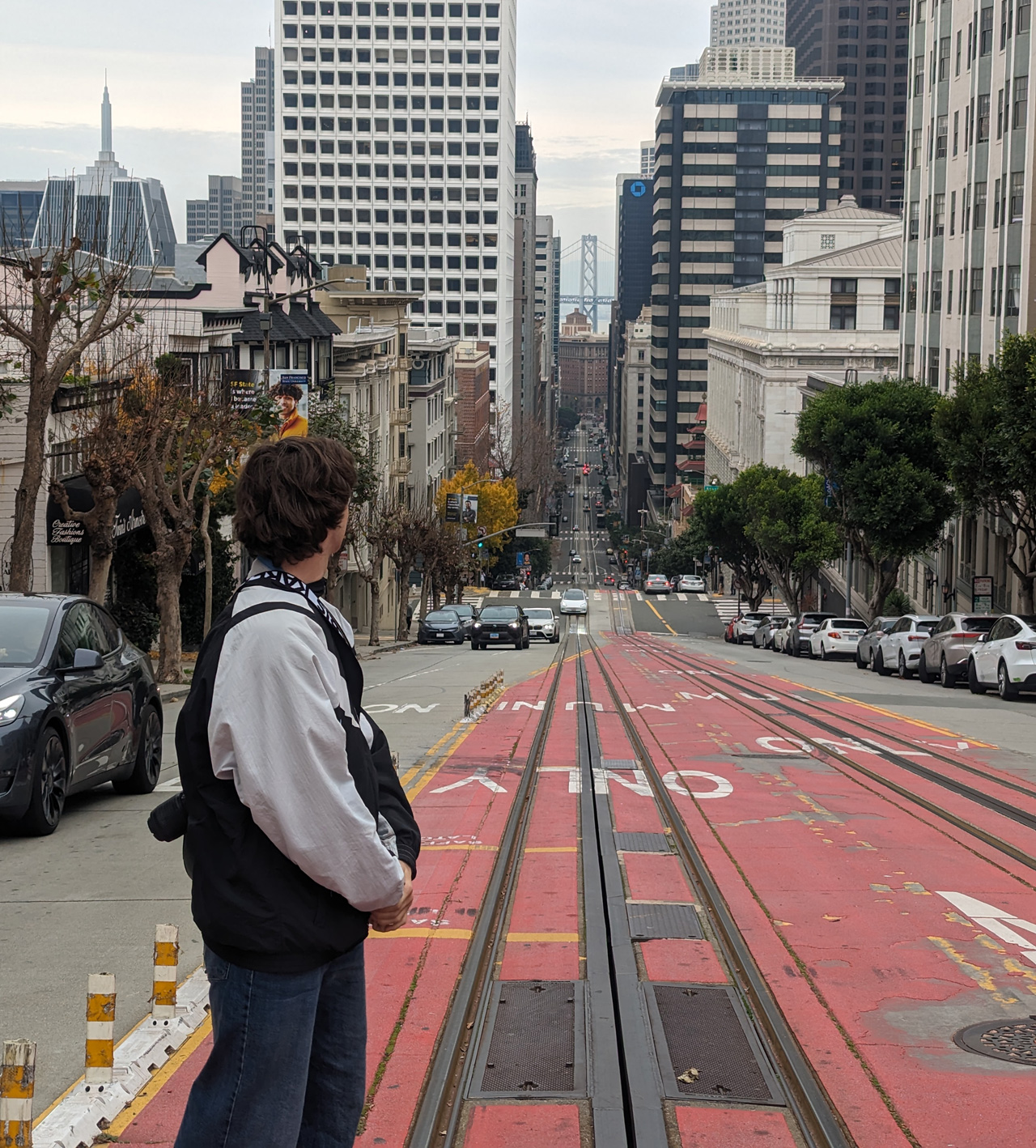 Cornelius von hinten. Vor ihm: Skyline von San Fran. Rote Straße, mit Schienen. 