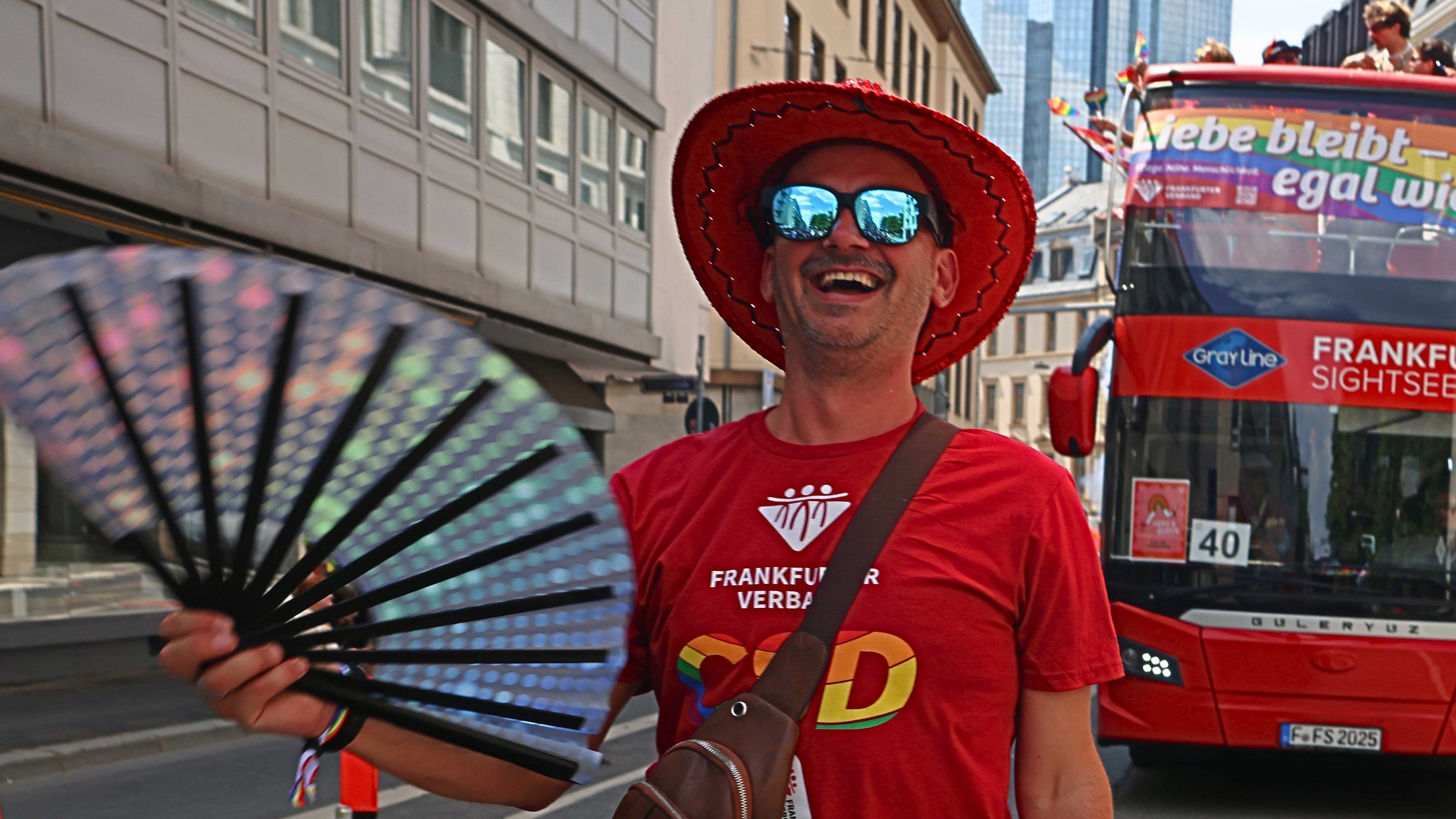 Ein Mann mit rotem Hut und Sonnenbrille lächelt und hält einen Fächer. Er steht auf einer Stadtstraße in der Nähe eines Sightseeing-Busses mit Regenbogen-Dekor.