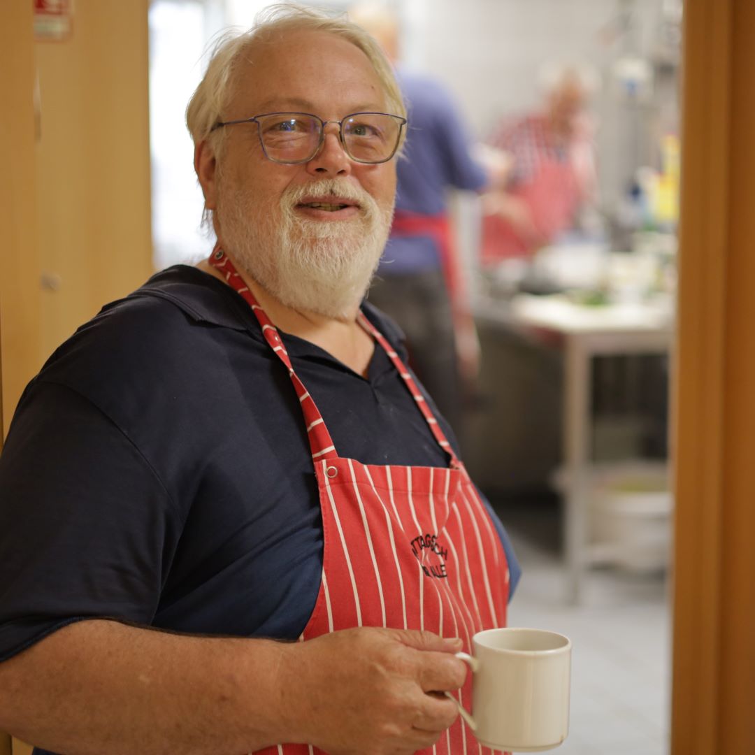 Kräftiger Mann mit weißem Bart, mit rotweiß-gestreifter Kochschürze und Kaffee
