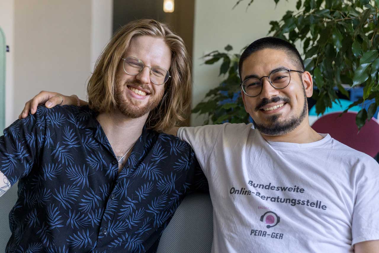 Kiril und Furkan sitzen auf einem Sofa Seite an Seite und lächeln in die Kamera.