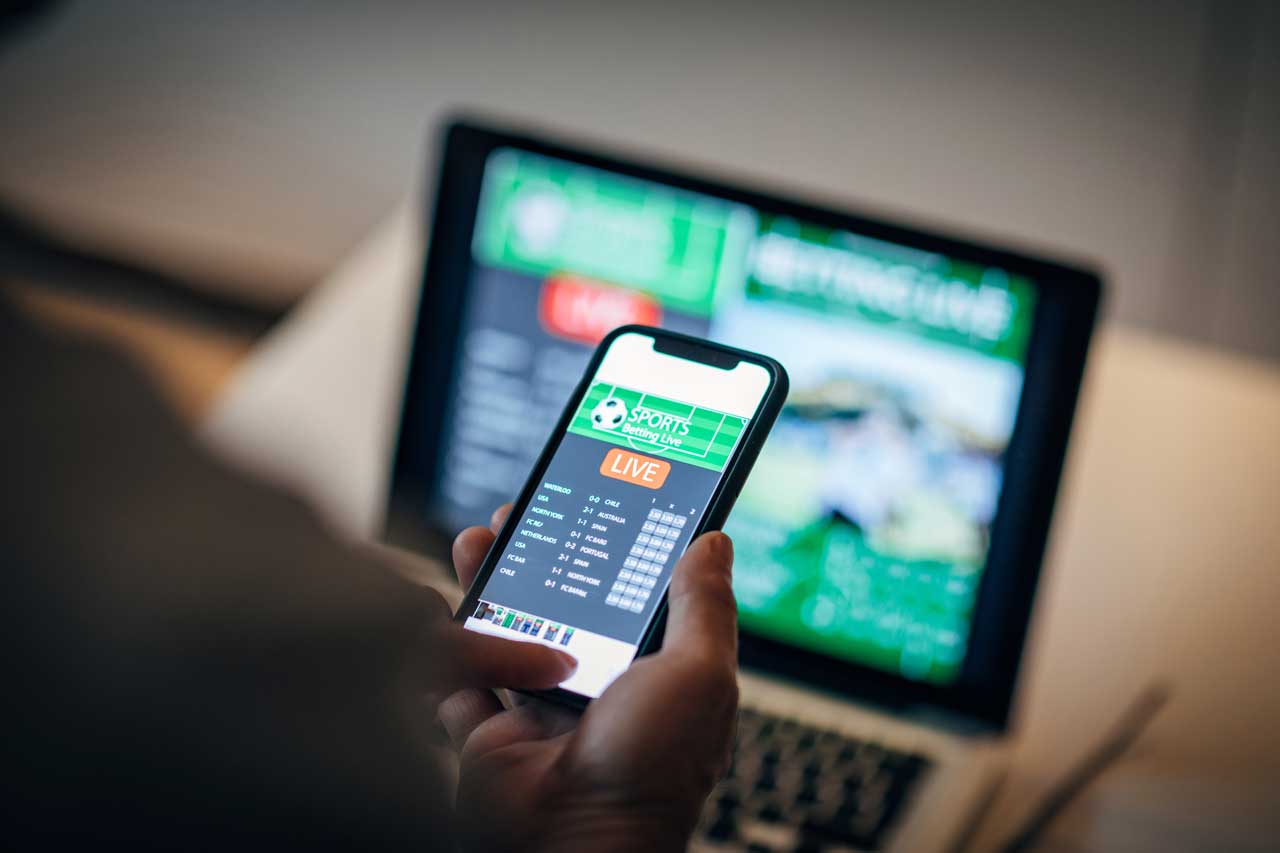 Eine Person hält ein Smartphone mit einer Live-Sportwetten-App in der Hand. Im Hintergrund ist ein unscharfer Laptop mit einer geöffneten Sportwetten-Website zu erkennen.