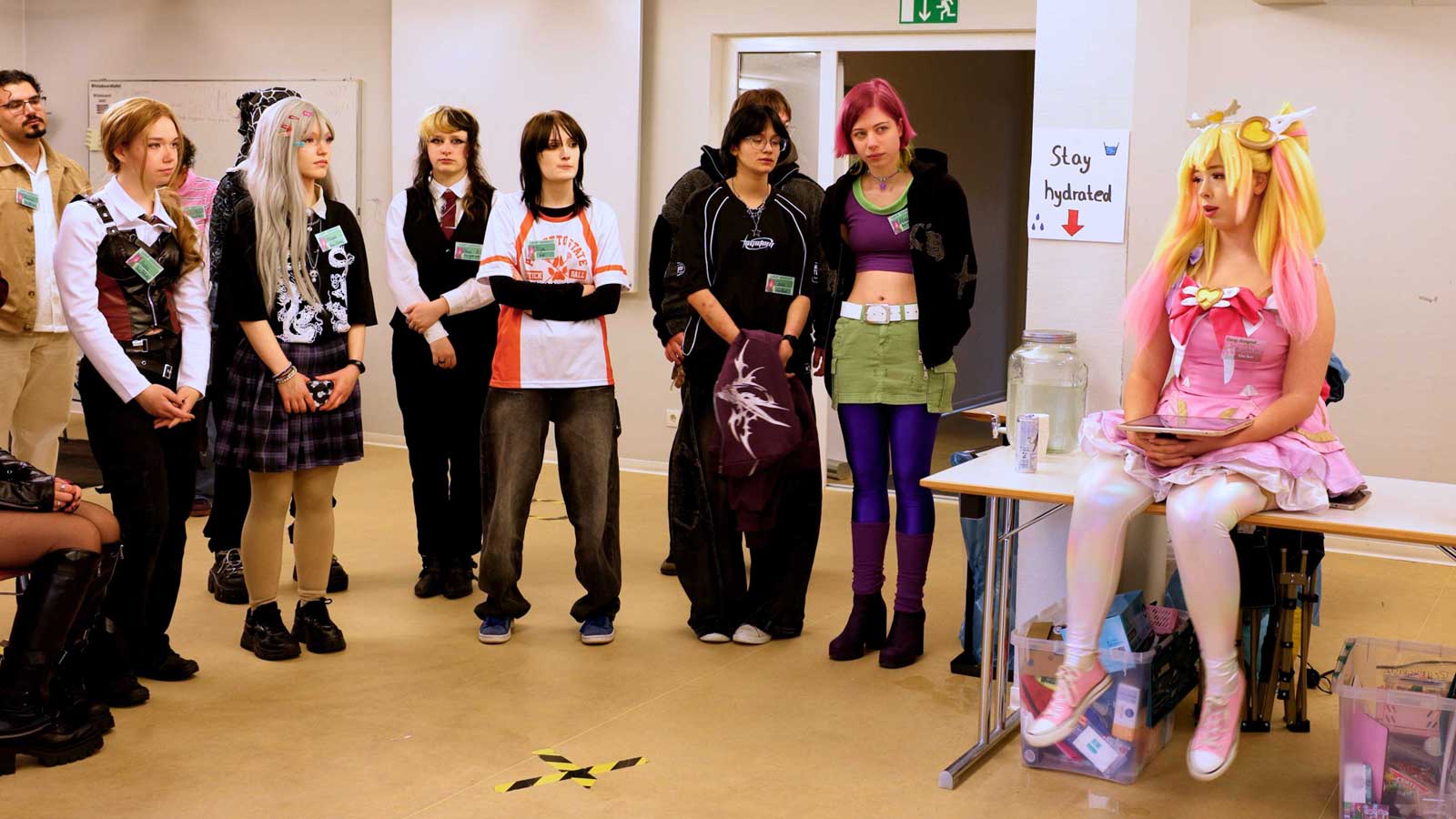 Gruppe von Menschen in unterschiedlichen Cosplay-Outfits steht in einem Innenraum vor einer Tür; rechts sitzt eine Person im auffälligen pink-gelben Kostüm an einem Tisch.