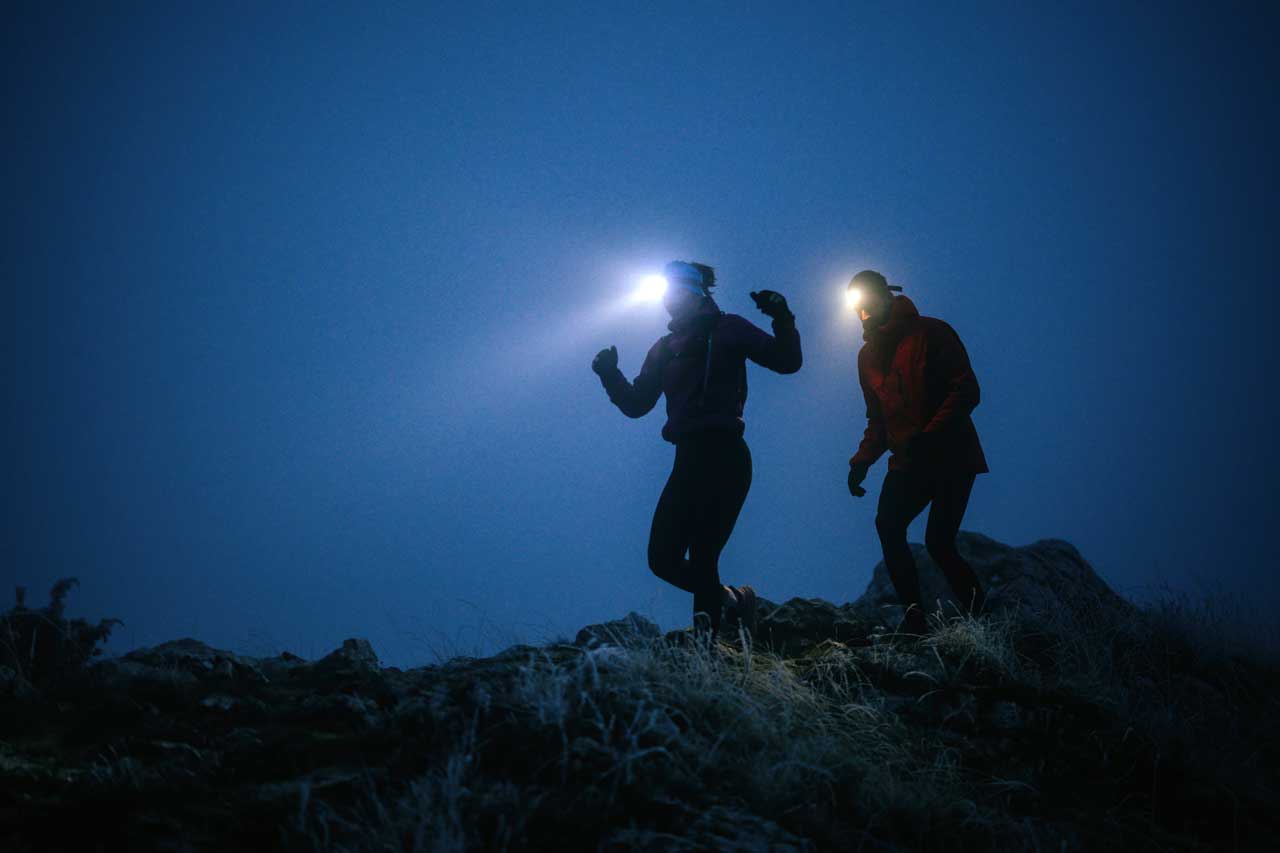 Zwei Personen wandern nachts über einen felsigen Weg und tragen Stirnlampen, deren Licht in die Dunkelheit strahlt.