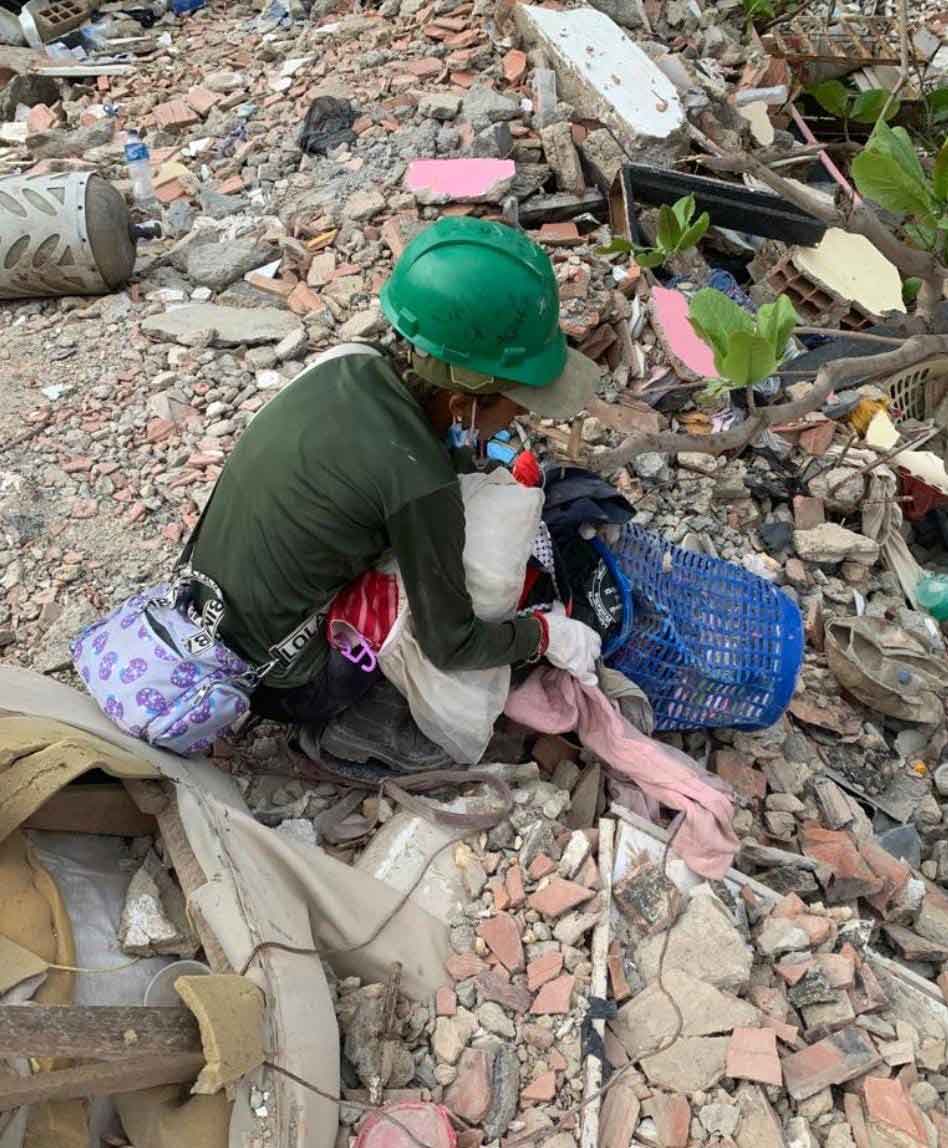 Eine Frau mit grünem Bauhelm hockt inmitten der Trümmer. Sie sammelt Habseligkeiten in einen blauen Wäschekorb aus Plastik.