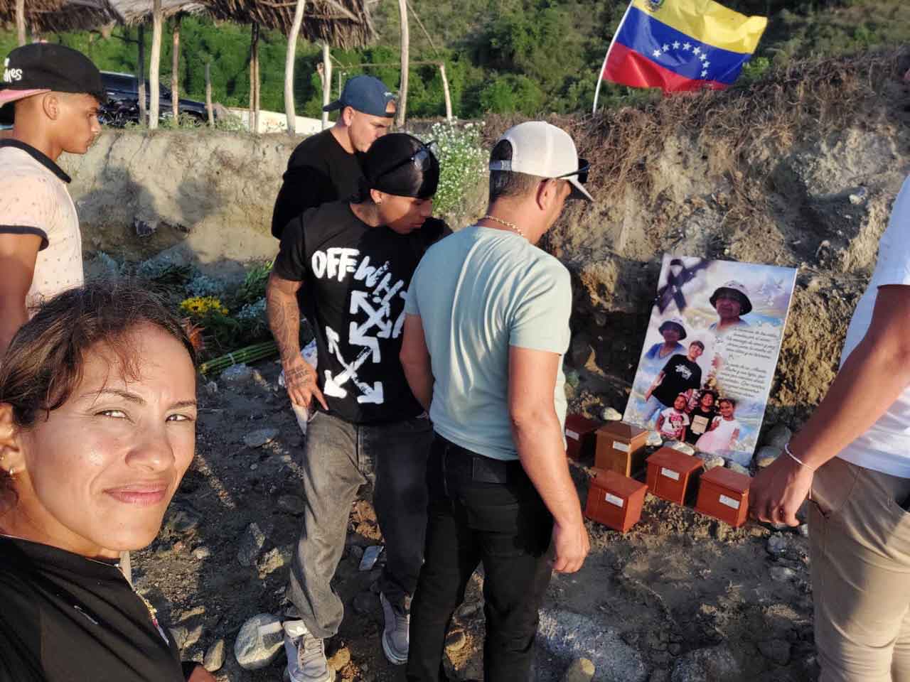 5 kleine Holzkästen stehen vor einem Plakat mit Bildern der verstorbenen Familie von von Gabriela. Im Hintergrund die Fahne Venezuelas, im Vordergrund Menschen, die diesen Menschen gedenken, eine Frau macht das Foto als Selfie. 