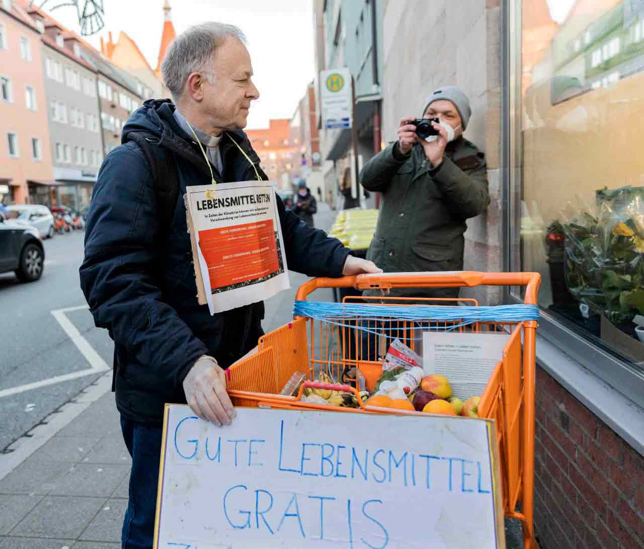 Jesuitenpater Jörg Alt steht mit einem Einkaufswagen voller Lebensmittel vor einem Schaufenster.