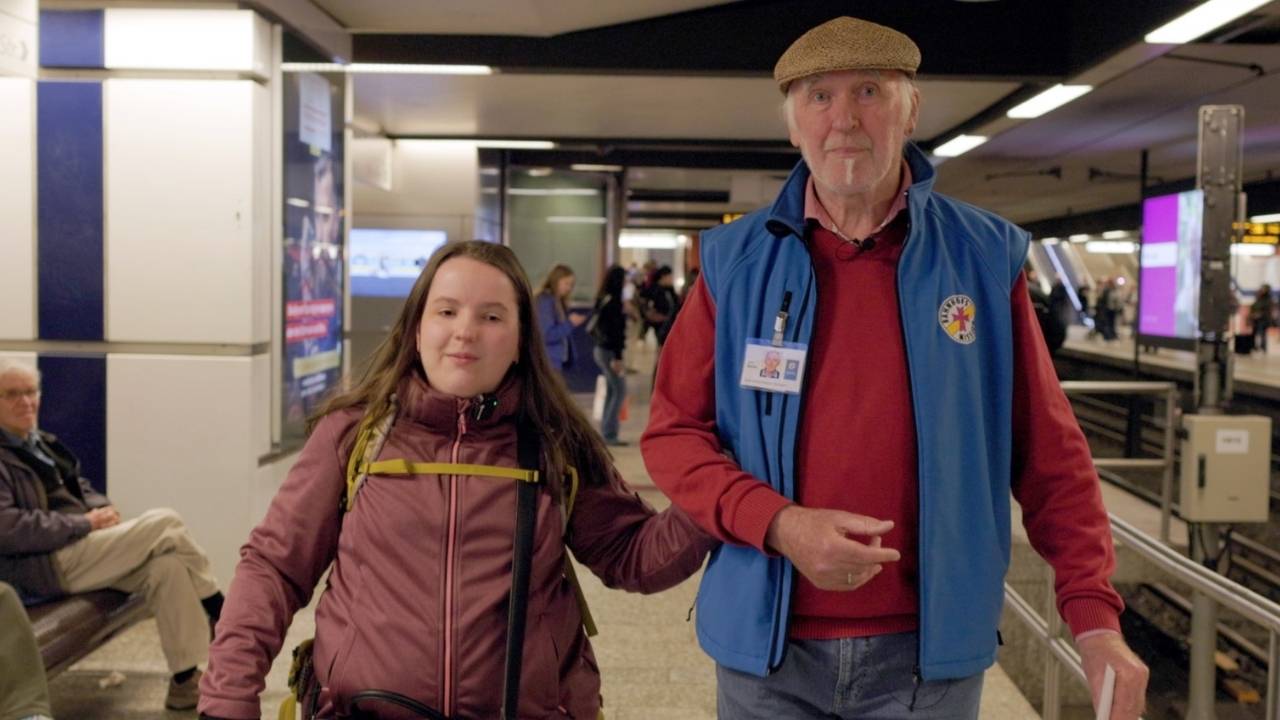 Die 21-jährige Olivia trägt dunkelrote Jacke und einen Rucksack. Sie hakt sich bei Josef ein. Der 74- Jährige trägt eine blaue Weste mit dem Logo der Bahnhofsmission