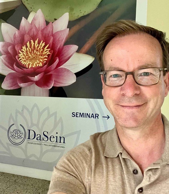 Das Bild zeigt Markus hinter einem Plakat, auf der eine rosa Seerose abgebildet ist und die Aufschrift "DaSein. Hospizdienst.Palliativberatung" zu lesen ist.