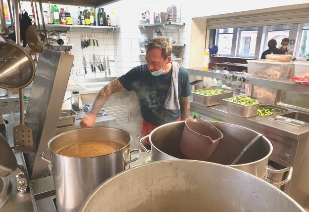 In einer großen Küche stehen einige dampfende Töpfe. Ein Koch der ada_kantine steht dahinter und rührt mit einer Kelle im Topf. Im Hintergrund sind Küchenutensilien und Gewürze.