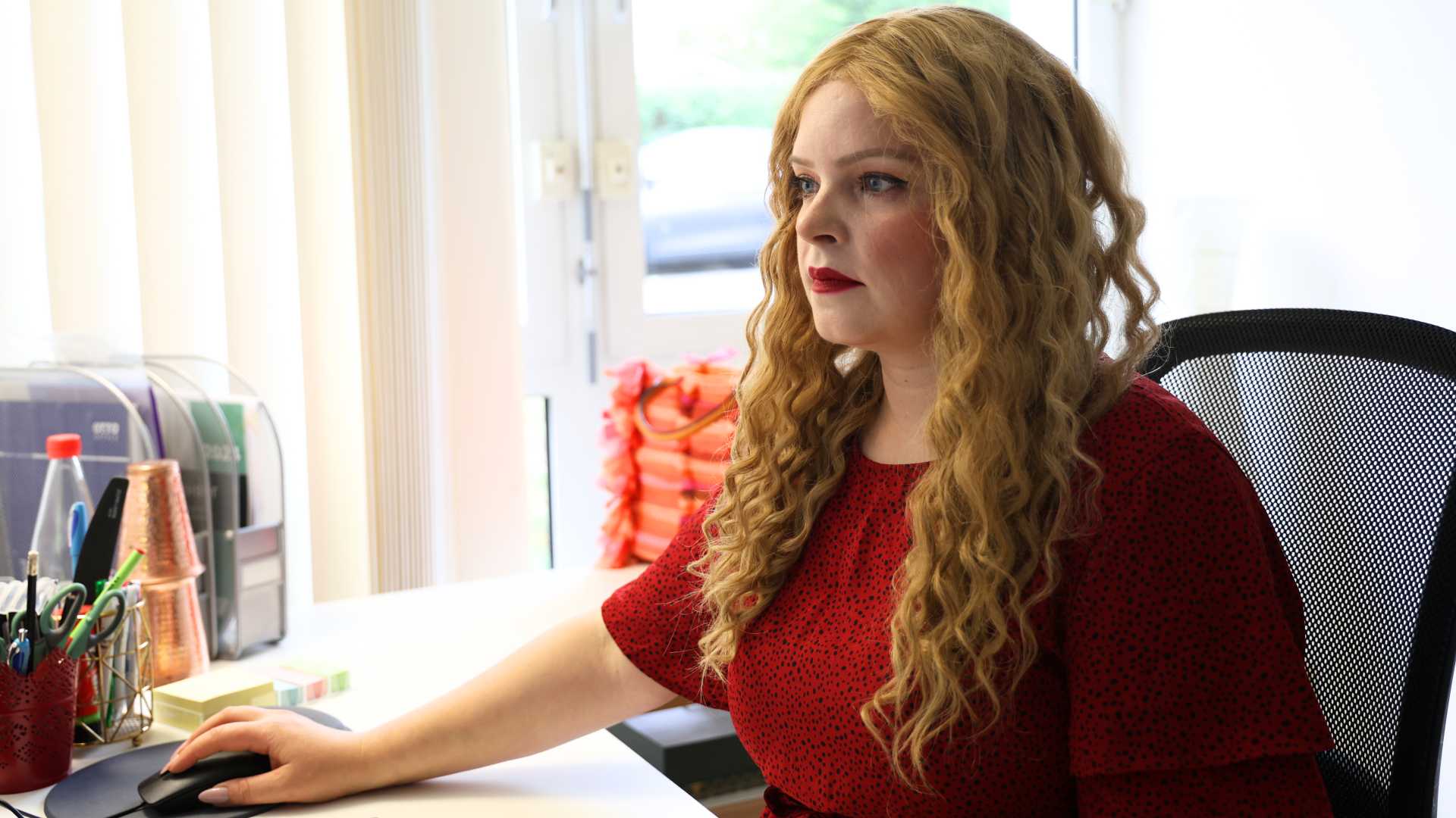 Juditz Desoi, eine Frau mit langen rotblonden Haaren. Sie trägt ein rotes Oberteil, roten Lippenstift und sitzt an ihrem Schreibtisch