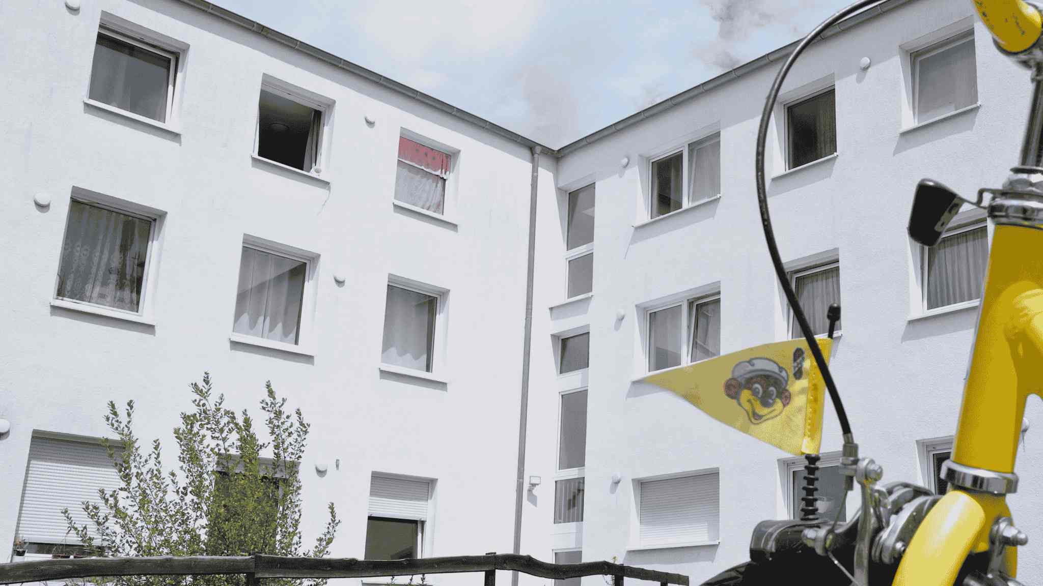 Zu sehen ist ein weißes, dreistöckiges Haus. Im Vordergrund ist ein Kinderrad angeschnitten zu sehen. 