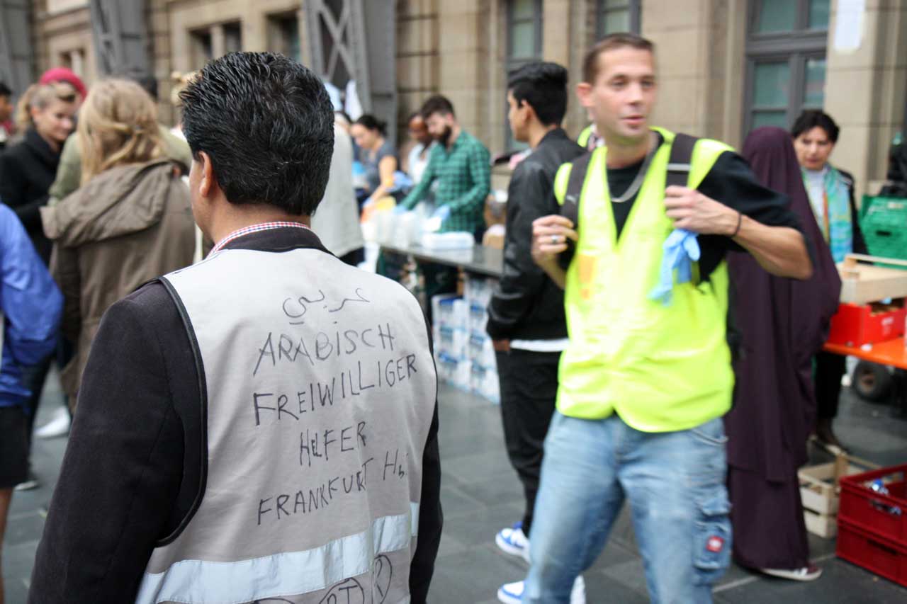 Ein Mann steht mit dem Rücken zur Kamera in einem Bahnhofsgebäude. Er trägt eine graue Weste, auf der in schwarzer Handschrift "ARABISCH FREIWILLIGER HELFER FRANKFURT HBF" steht. Im Hintergrund sind weitere Menschen, die sich um Tische mit Hilfsgütern versammelt haben. Ein Mann in einer neongelben Weste blickt nach vorne, während er sich durch die Menschenmenge bewegt.