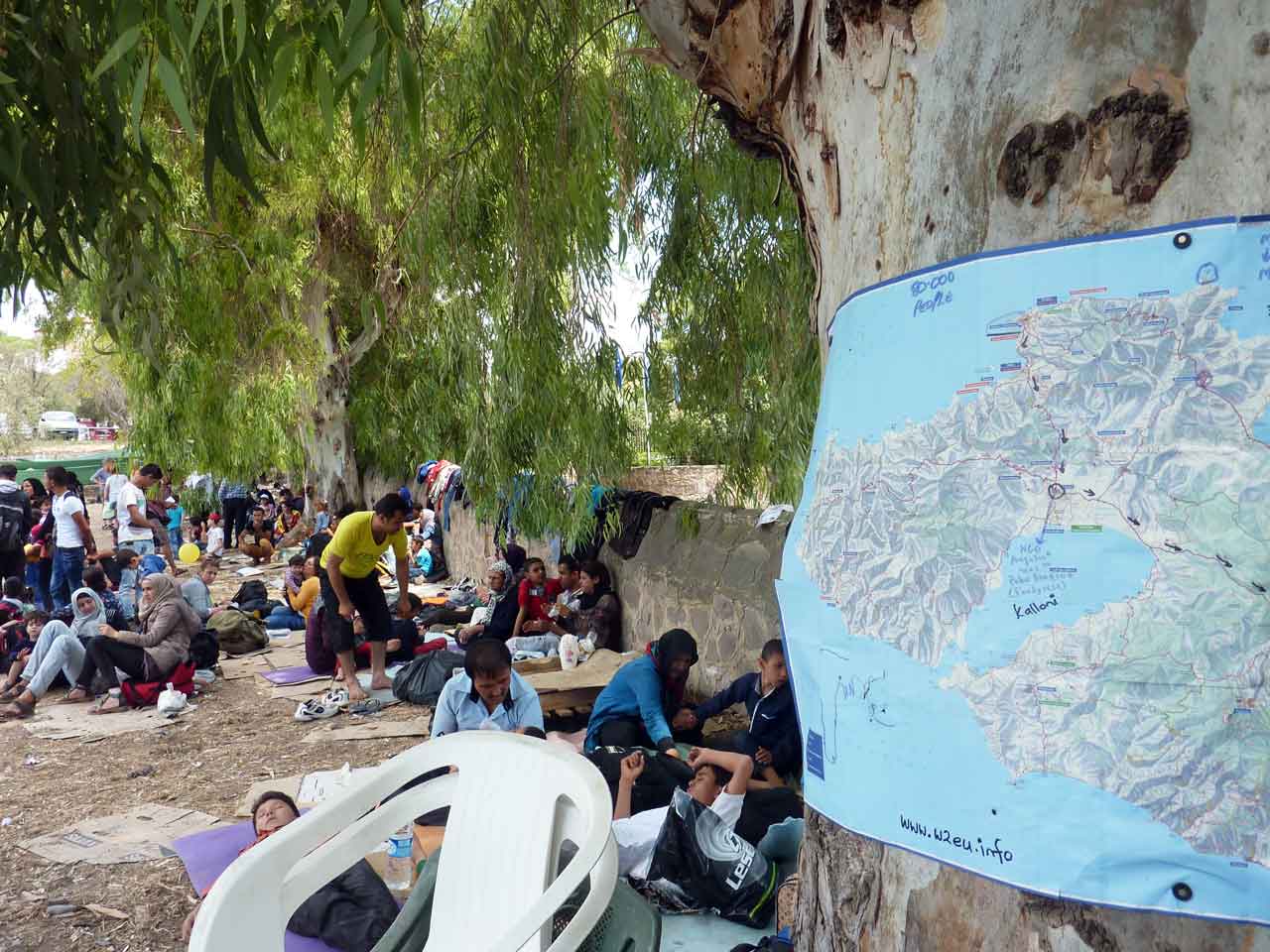 Eine Gruppe von Menschen liegt oder sitzt im Schatten von Bäumen und einer Mauer. Im Vordergrund hängt eine Karte, die die Insel Lesbos und die umliegenden Gewässer zeigt. Im Bildhintergrund sind mehr Menschen zu sehen.