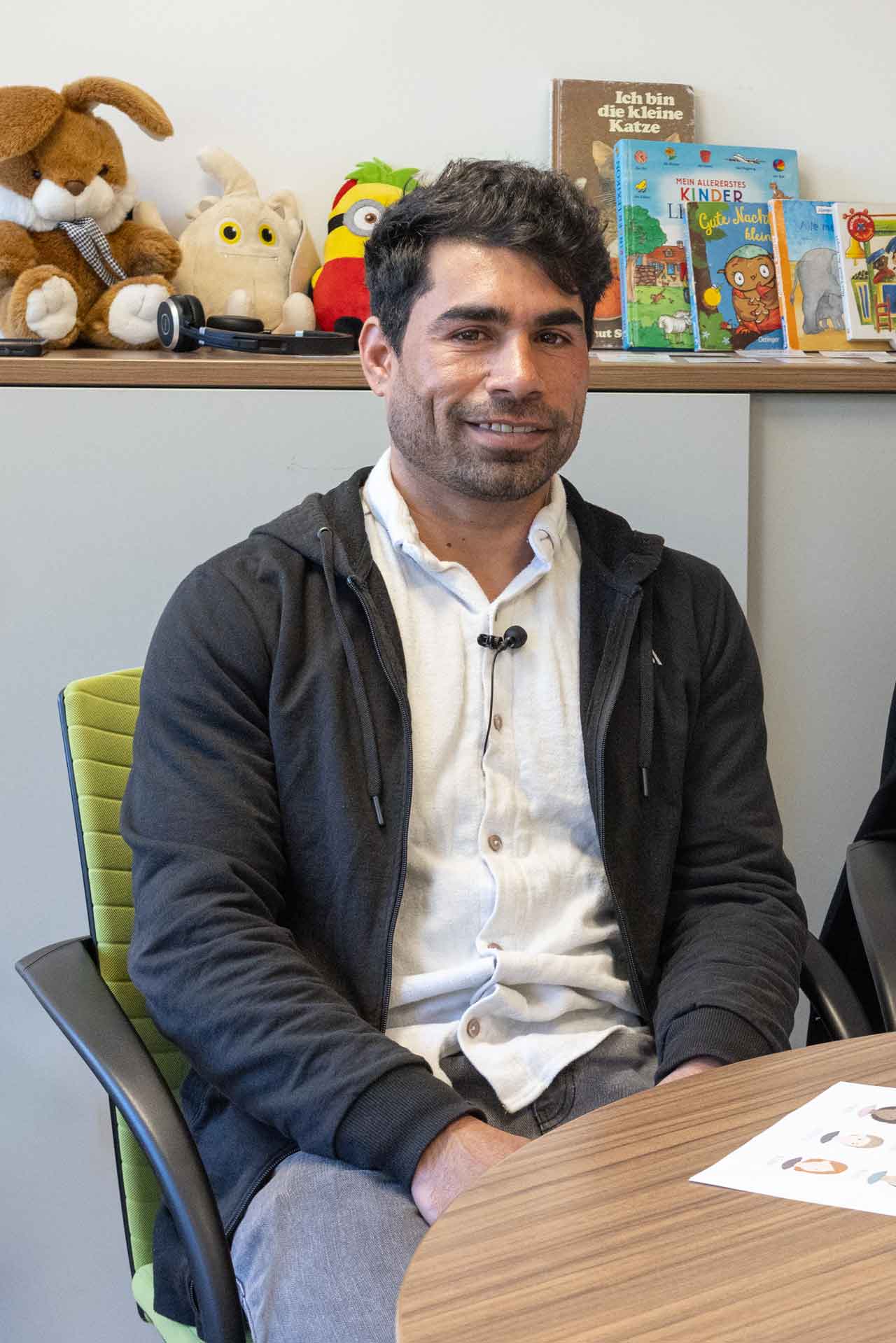 Jamshid Qaderi sitzt an einem Tisch im Evangelischen Zentrum am Weißen Stein. Auf dem Schrank hinter ihm sind Kuscheltiere und Kinderbücher.