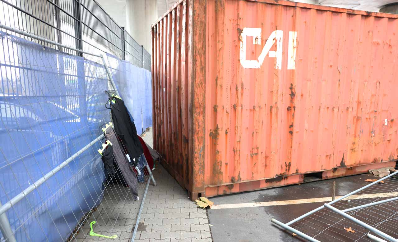 Orangener großer Container, Bauzäune mit Kleidung auf Kleiderbügeln. 