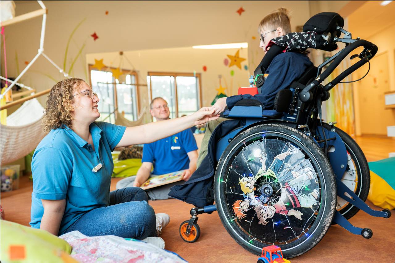Eine Pflegerin und ein Pfleger befinden sich im einem Jungen im Rollstuhl in einem Spielraum in der Familienherberge