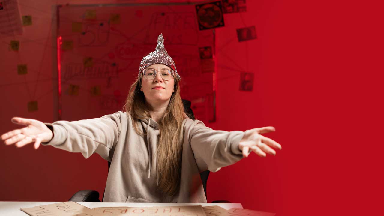 Eine junge Frau mit langen blonden Haaren sitzt mit Hoodie, Brille und Alu-Hut an einem Tisch. Sie macht eine große Geste mit offenen Armen. Ihr Gesichtsausdruck wirkt überlegen und allwissend.  