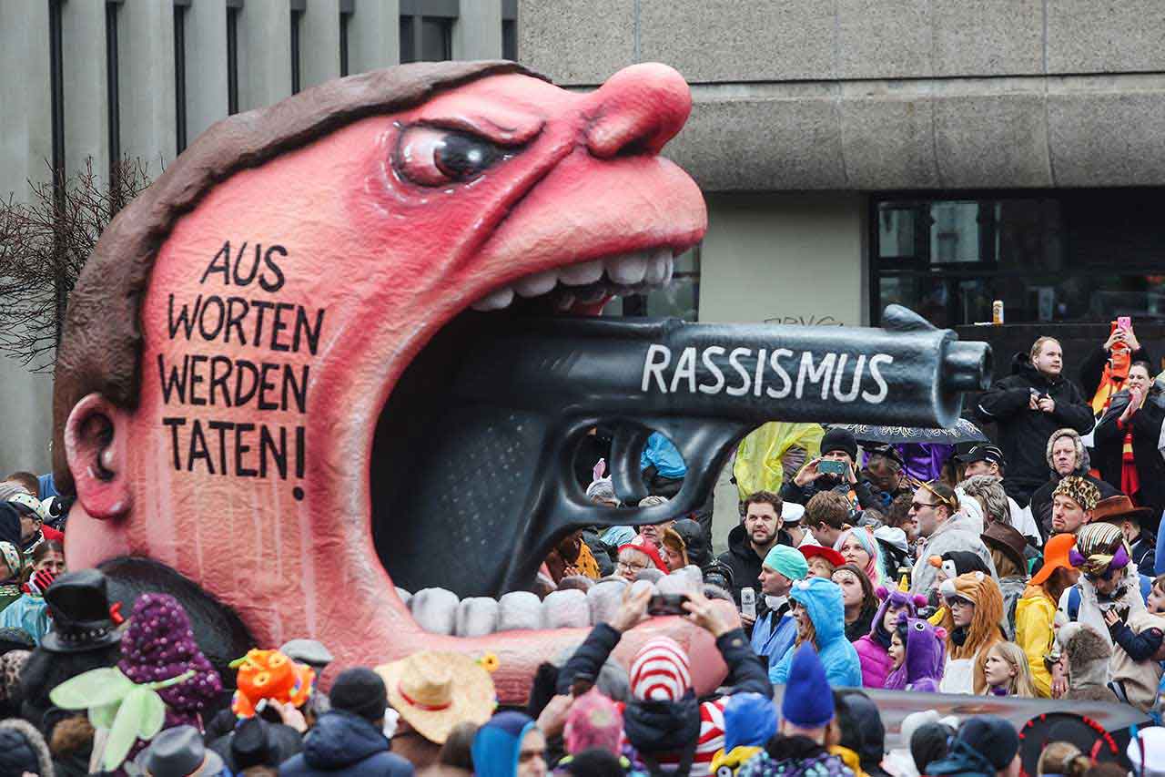 Mottowagen beim Düsseldorfer Rosenmontagszug 2020. Er trägt die Aufschrift: "Aus Worten werden Taten!"  Aus dem Mund eines wütend blickenden Kopfes ragt eine Waffe, auf der "Rassismus" steht.