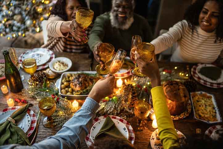 Familie sitzt um voll gedeckten Essenstisch, Weihnachtsbaum im Hintergrund, alle stoßen an.