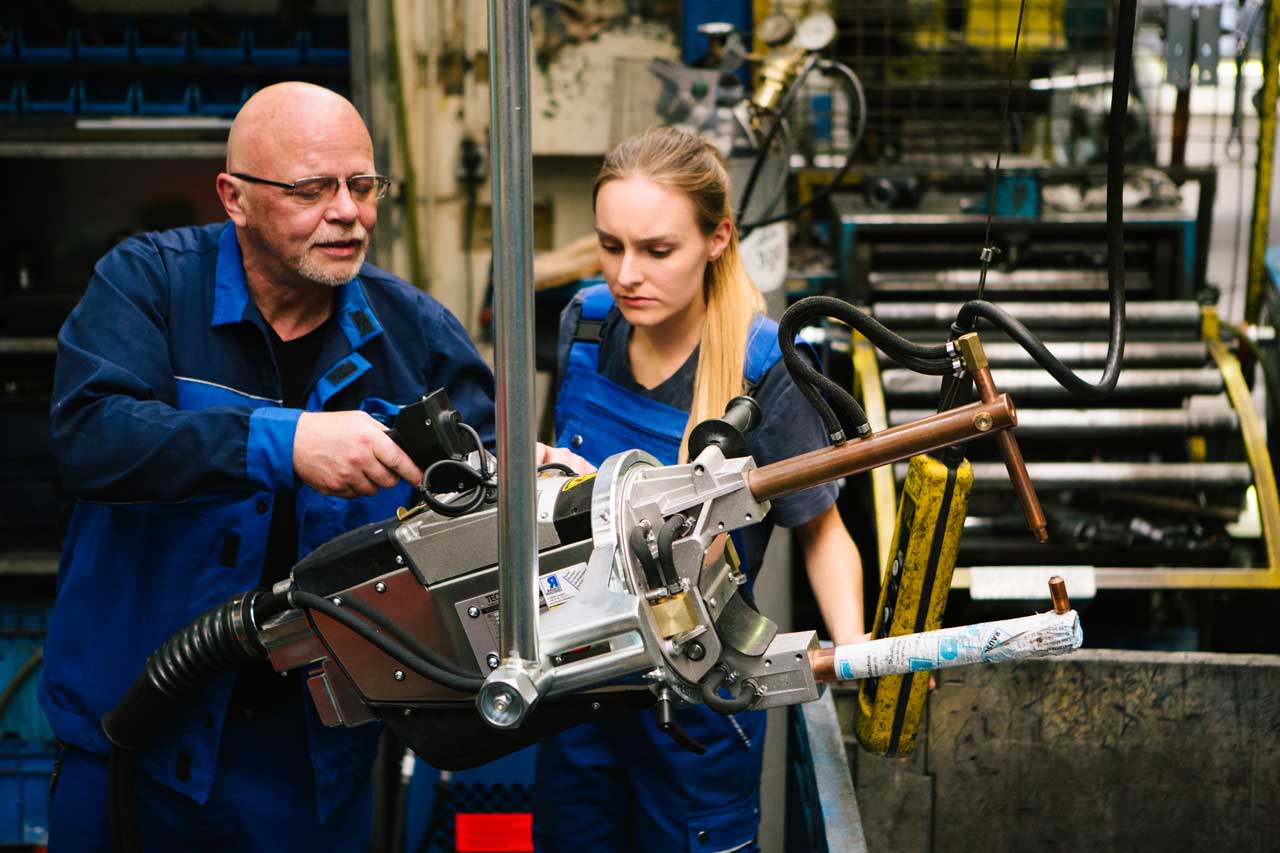 Ein erfahrener und älterer männlicher Mitarbeiter gibt der weiblichen Auszubildenden Anweisungen in der Metallindustrie