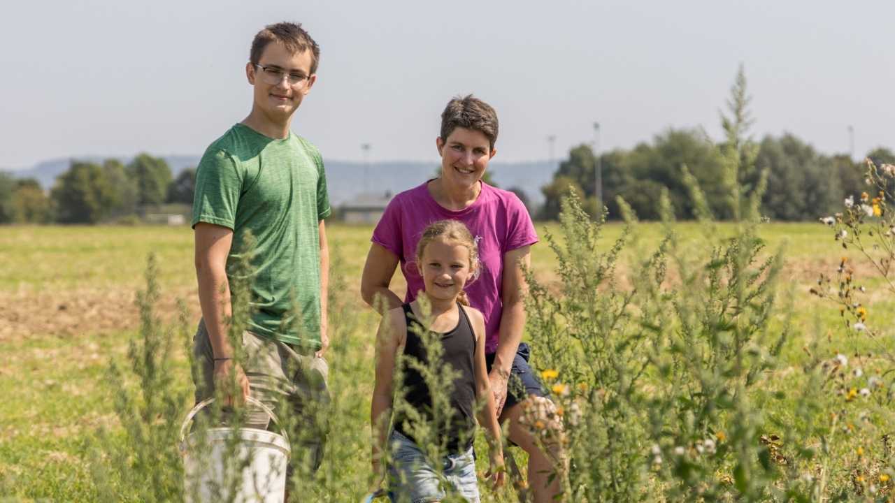 Ein Jugendlicher im grünen T-Shirt steht neben einer Frau und deren Tochter auf einer grünen Sommerwiese. Alexander hält einen weißen Eimer in der Hand.