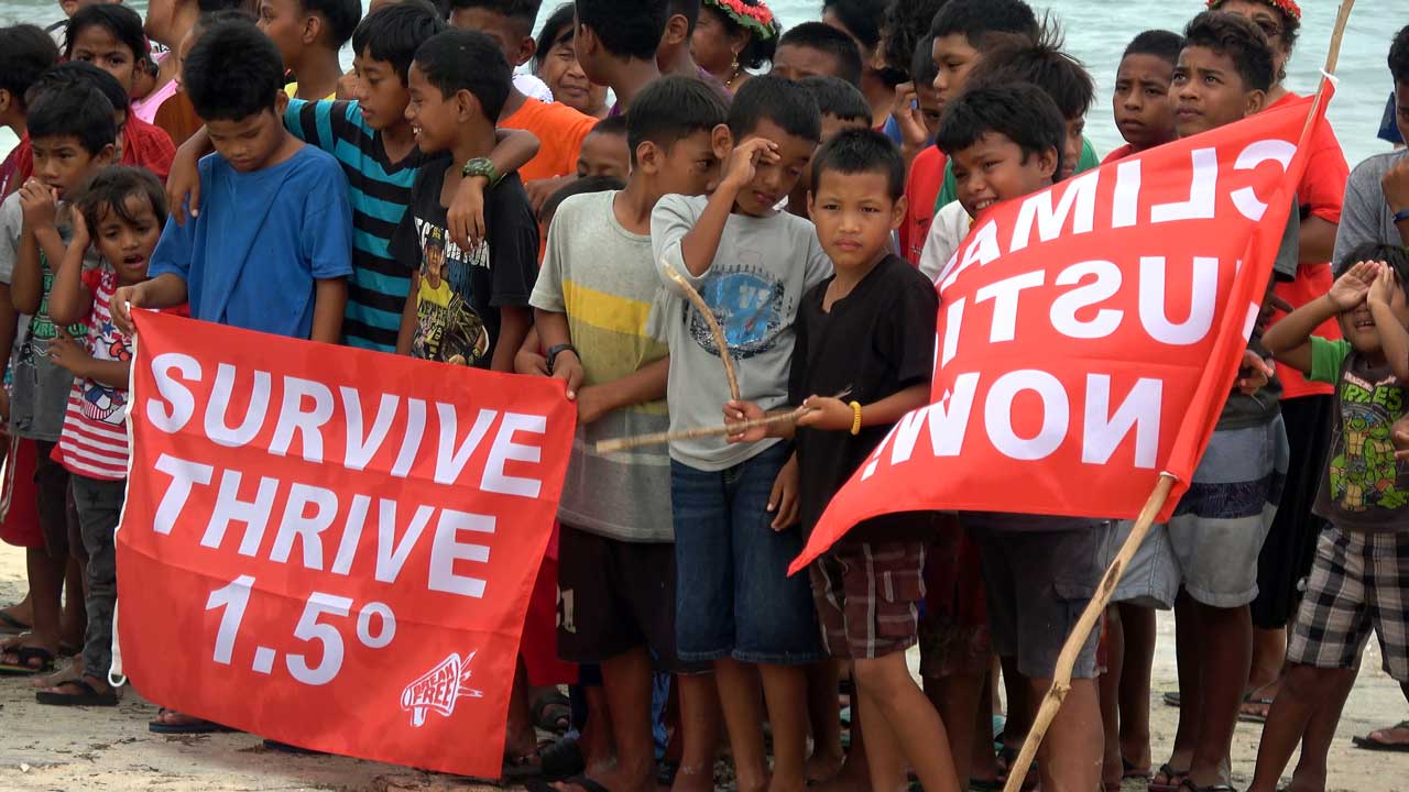 Kindergruppe hält rote Banner mit der Aufschrift „Survive Thrive 1.5º“ und „Climate Justice Now“ bei einer Klimaprotestveranstaltung am Strand.