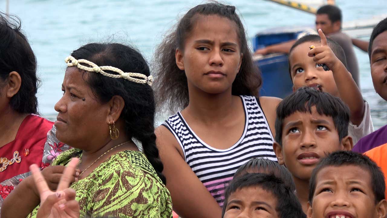 Eine Gruppe von Menschen steht am Wasser. Eine Person trägt ein rotes, mit Blumen besticktes Kleid und goldene Ohrringe. Im Hintergrund sind ein Boot und das Meer zu sehen.
