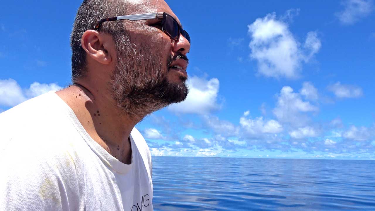 Ein Mann in einem weißen T-Shirt steht vor einem klaren Ozean und einem blauen Himmel mit vereinzelten Wolken.