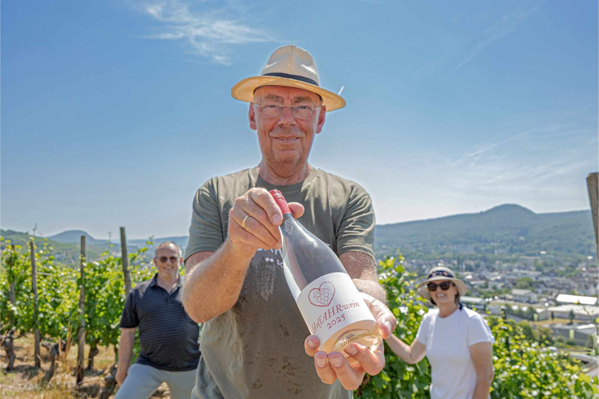 Ein Mann mit Strohut steht in einem Weinberg. Im Hintergrund das Ahrtal und zwei weitere Menschen unscharf. Der Mann hält eine Weinflasche in die Kamera mit der Aufschrift „HelfAHRwein“