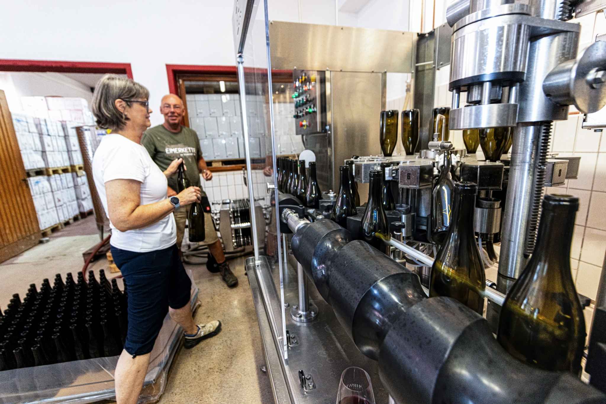 Zwei Personen stehen vor einer Maschine, die Wein in Weinflaschen abfüllt. Eine Frau mit schwarzer Hose und weißen Shirt und eine Mann mit grünem Shirt