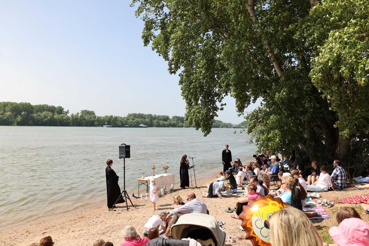 Besucher sitzen am Rheinstrand und schauen einer Taufe zu.