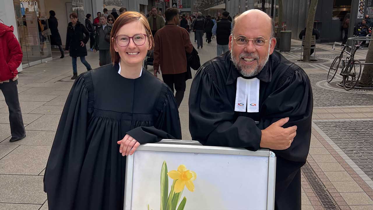 Vikarin und Pfarrer stützen sich auf ein Schild, auf dem „Ostersegen“ steht.