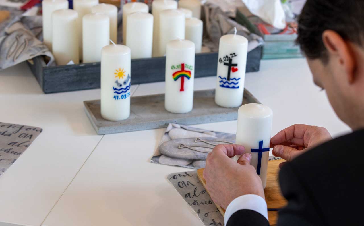 Mehrere weiße Kerzen. Ein Mann beklebt gerade eine Kerze mit einem blauen Kreuz.
