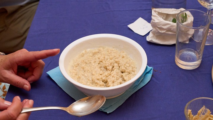 Eine kleine Schüssel Porridge auf einer blauen Tischdecke. Davor zwei Hände, eine hält einen Löffel. Daneben Tischdeko und ein Wasserglas
