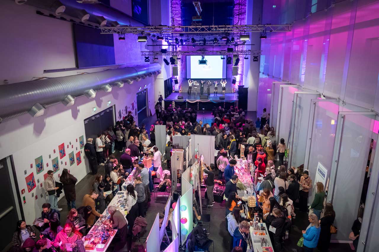 Junge Frauen tanzen auf der Bühne (im Hintergrund) beim Korea Culture Day 2025 in der Jugendkulturkirche Sankt Peter in Frankfurt am Main am 25.10.2025. Im Vordergrund sind diverse Stände. 