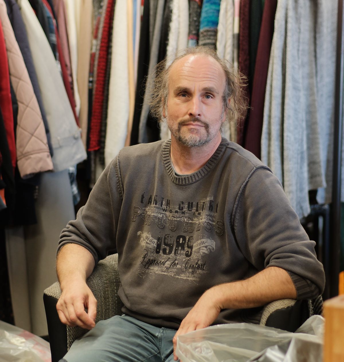 Ein Mann mit lichtem Haar und Schnäuzer sitzt auf einem Stuhl hinter ihm sind Kleiderstangen mit Jacken und Mäntel zu erkennen.