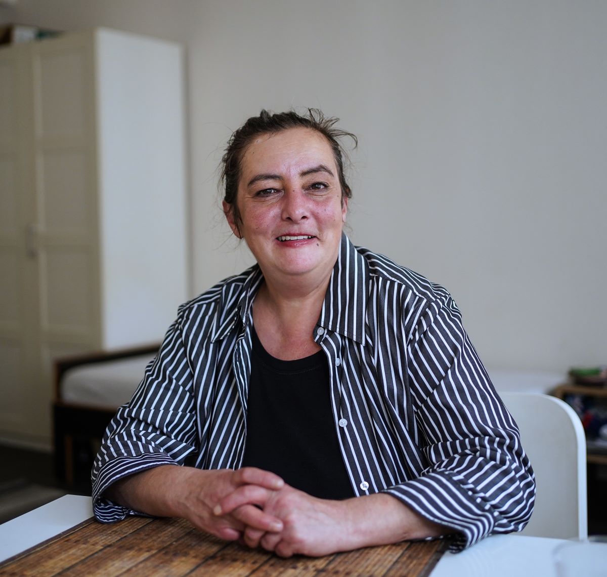 Eine dunkelhaarige Frau sitzt an einem Tisch, die Hände vor sich auf dem Tisch einander verschränkt und lächelt in die Kamera.